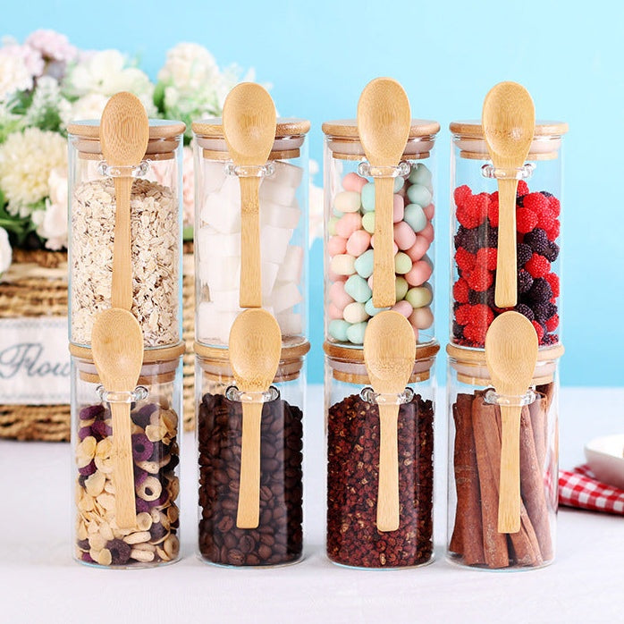 Bocaux en verre transparents avec cuillères en bois, contenant céréales, sucre, confiseries, café, épices et bâtons de cannelle, pour rangement esthétique et fonctionnel en cuisine.