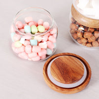 Récipient en verre transparent avec couvercle en bois d'acacia contenant des guimauves colorées, à côté d'un bocal similaire contenant des noix et muni d'un couvercle en bois, posé sur une table.