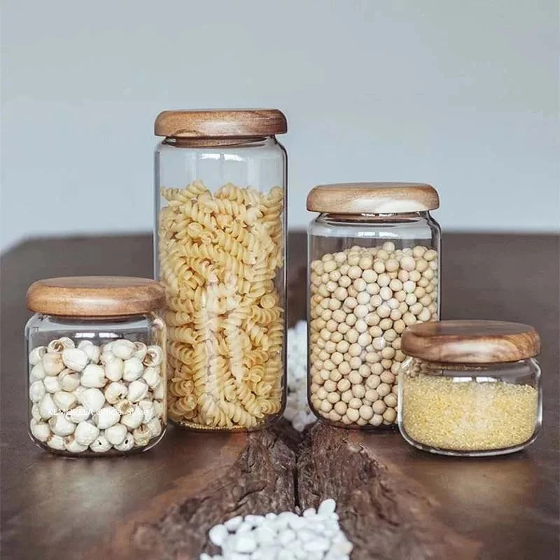 Set of transparent glass food storage containers with round acacia wood lids, shown holding pasta, beans, and grains on a wooden surface