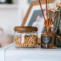 Récipient de conservation alimentaire avec corps en verre transparent et couvercle en bois d'acacia rempli de granulés alimentaires ronds beiges, posé sur une surface blanche à côté d'un flacon diffuseur en roseau brun.